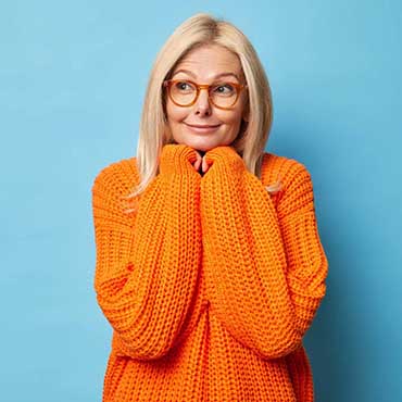 Beautiful blonde thoughtful middle aged woman keeps hands under chin and thinks deeply about something looks aside pensively wears spectacles knitted orange jumper isolated over blue studio wall