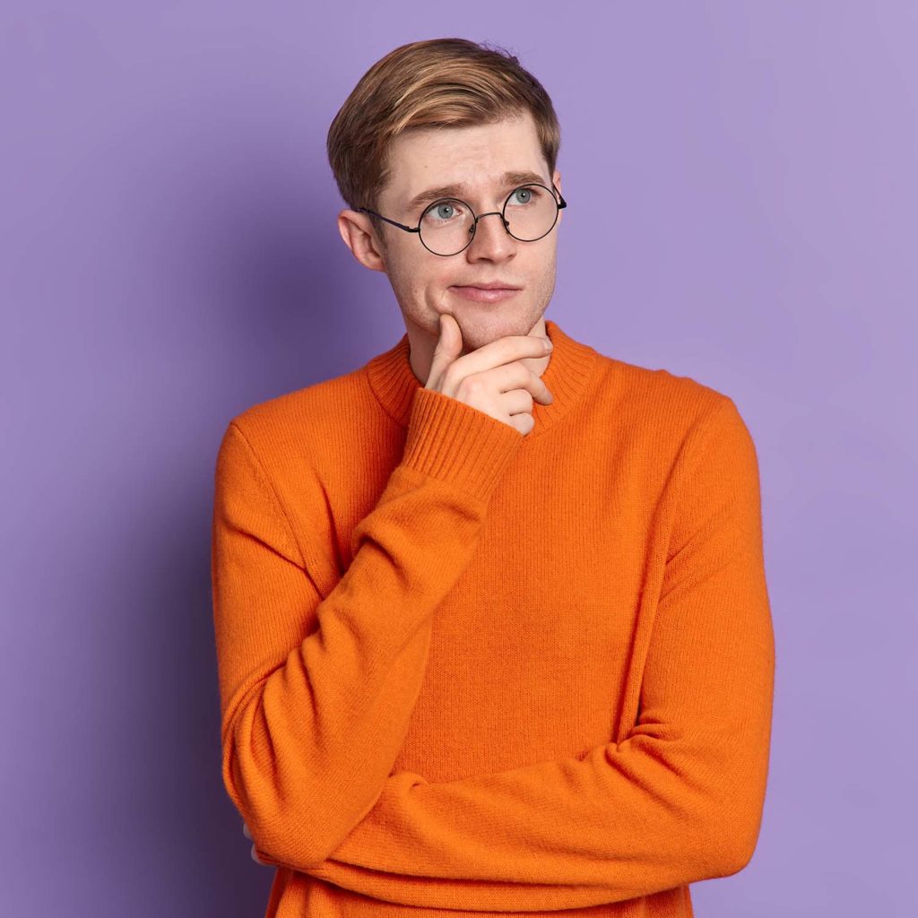 Portrait of blue eyed guy ponders on something stands in thoughtful pose holds chin concentrated into distance wears casual orange jumper isolated over purple background. Let me think a bit.